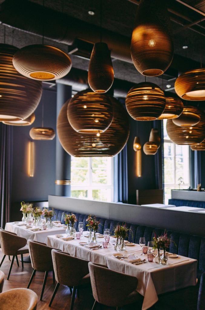 Festively set dinner area in the Mbassy by Franks restaurant at The Wellem Düsseldorf with elegant table decorations, velvety blue banquette and artistic, warmly lit designer lamps.
