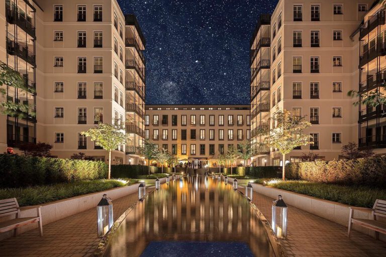 Illuminated inner courtyard of an elegant residential and hotel ensemble with symmetrical facades, water basins, lanterns and a starry night sky.