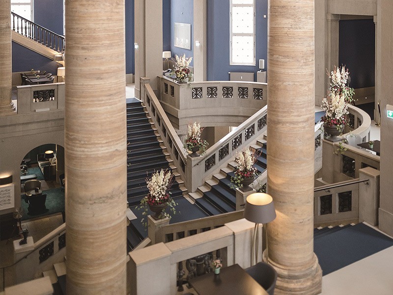 Historic staircase with decorative floral arrangements and tall columns in the elegant entrance hall of Hotel The Wellem Düsseldorf.