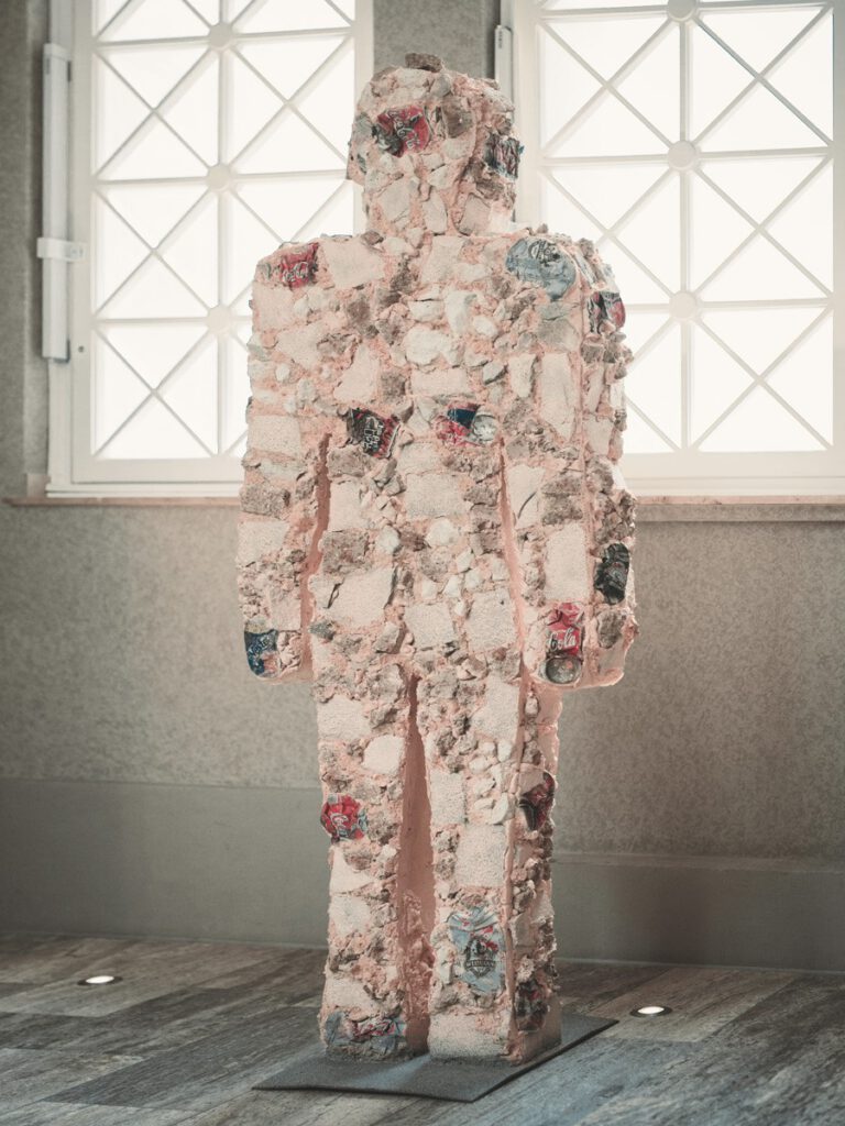 Contemporary figure sculpture at Hotel The Wellem Düsseldorf, made from structured fragments and incorporated can reliefs, presented in front of large geometric window surfaces.