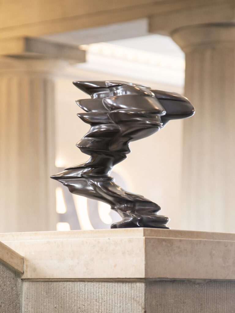 Moderne Skulptur in der Eingangshalle des Hotel The Wellem Düsseldorf, bestehend aus dynamisch geformtem, glänzendem Metall und präsentiert auf einem eleganten Steinpodest.