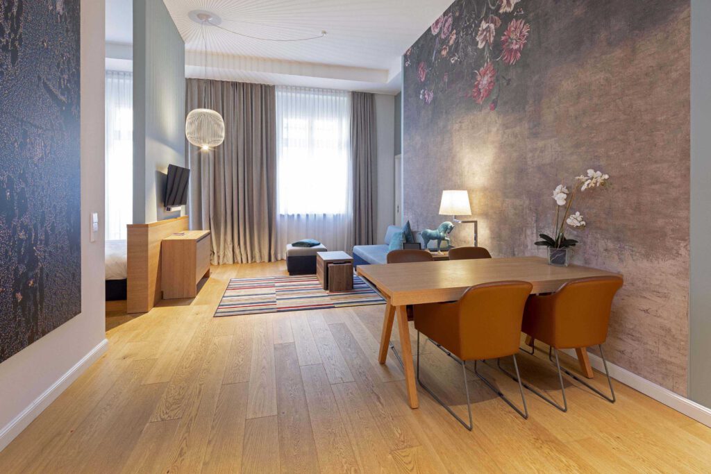 Elegant executive suite at Hotel The Wellem Düsseldorf with large living area, wooden dining area, stylish floral accent wall, designer sofa and separate sleeping area.