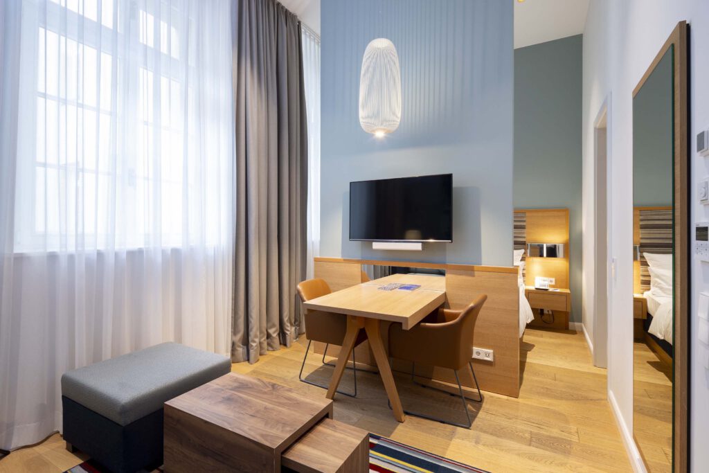 Modern studio at Hotel The Wellem Düsseldorf with desk, TV, warm wooden accents and view into the bright sleeping area with comfortable double bed.