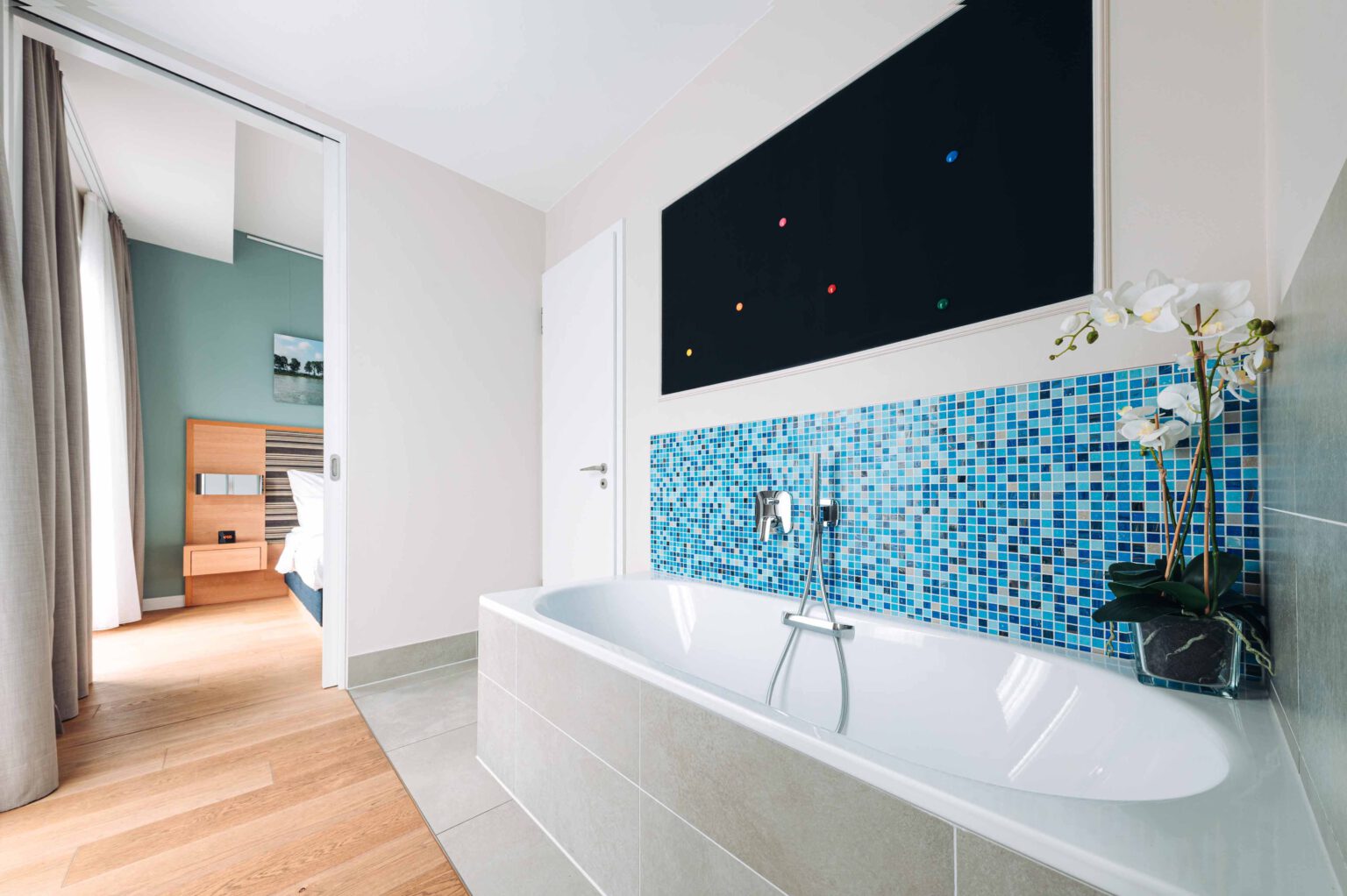 Modernes Badezimmer der Studio Suite mit eleganter Badewanne, blauen Mosaikfliesen, dezenter Wandkunst und Blick ins helle Schlafzimmer.