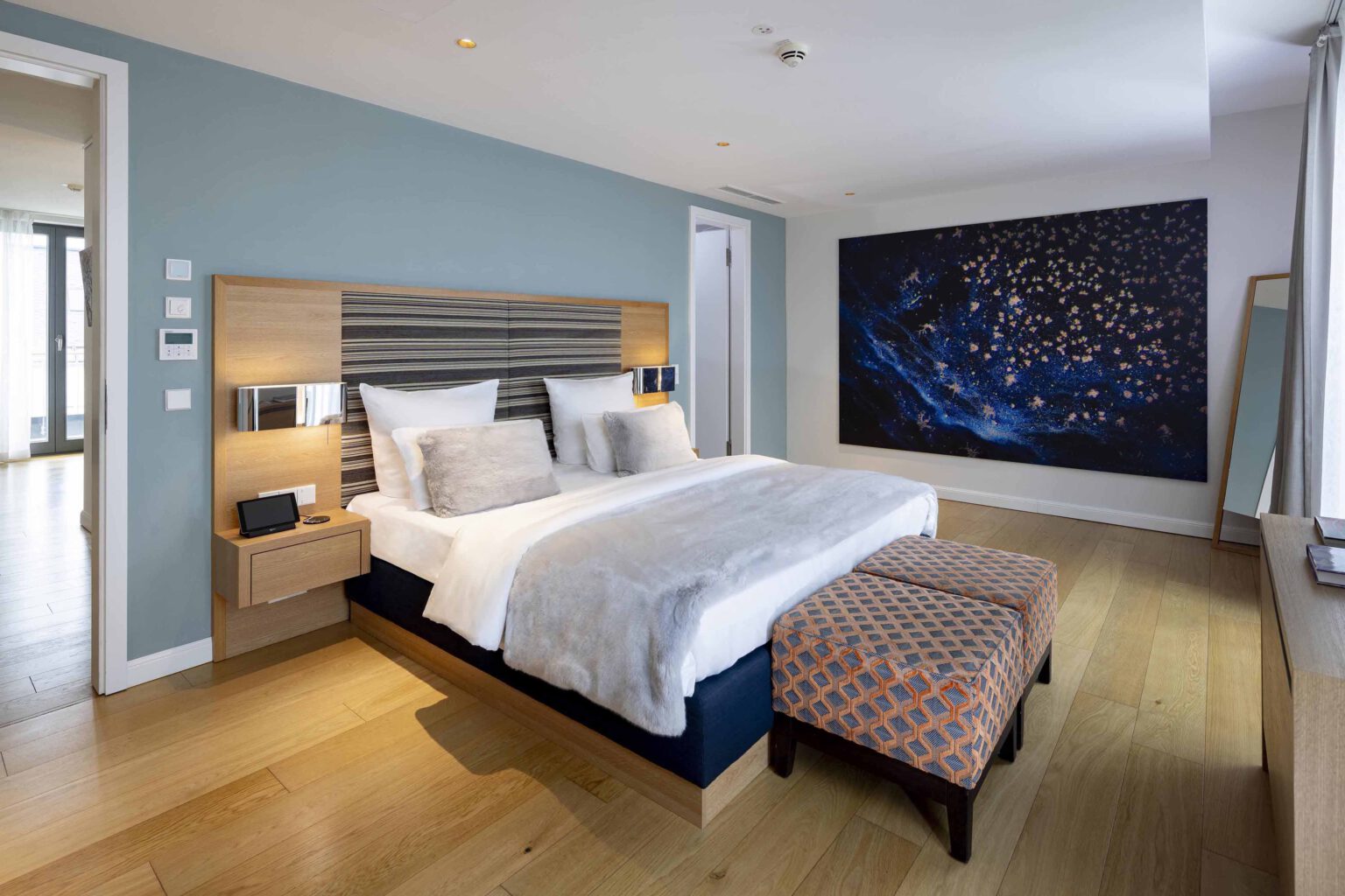 Spacious bedroom in The Wellem Suite with modern king-size bed, elegant wooden furnishings, light blue wall accents and large-scale artwork in the luxurious hotel The Wellem in Düsseldorf.