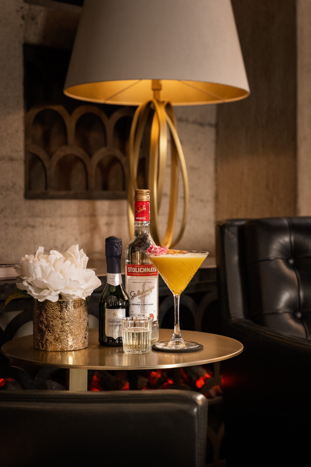 Stylishly arranged cocktail with vodka bottle, champagne mini and water glass on a golden side table in the elegant lounge of The Wellem Bar.