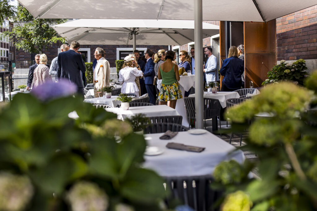 Sommerliches Get-together auf der Terrasse des Restaurants im Hotel The Wellem Düsseldorf mit elegant gekleideten Gästen unter großen Sonnenschirmen.